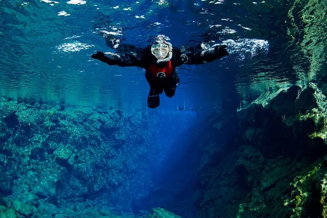 A snorkeler in a drysuit seen from below, arms spread, above the blue fissure